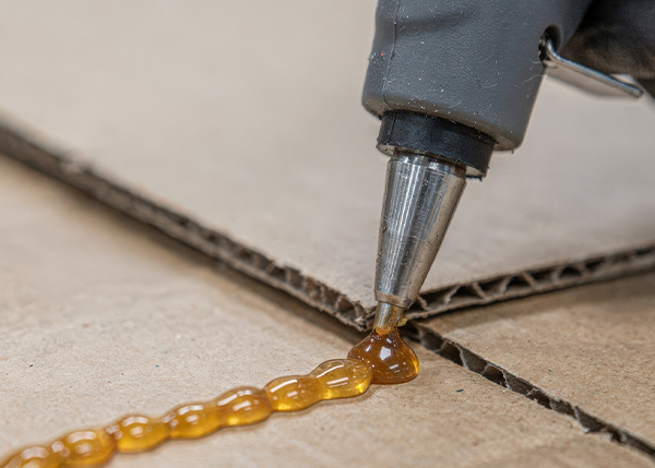 Nordson-compatible applicator gun dispensing adhesive on cardboard surface.